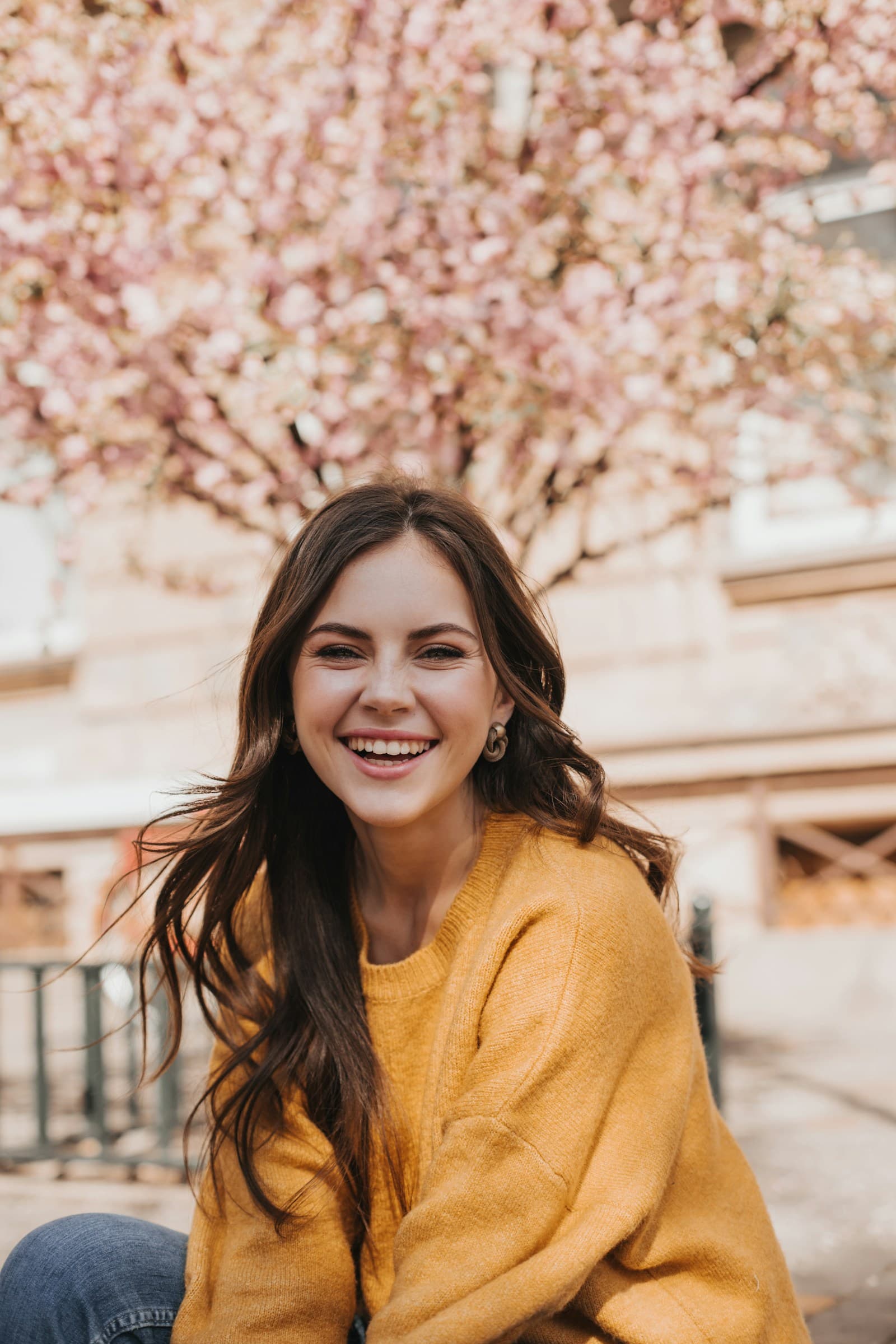 A smiling woman in a mustard jumper outdoors, with a flowering tree behind her.